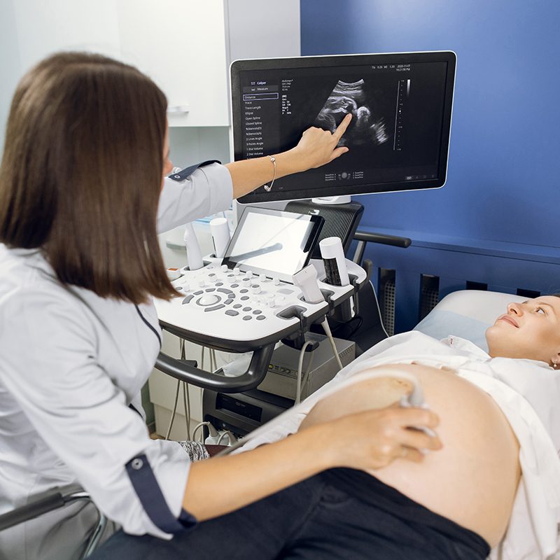 Back view of female ultrasound technician, scanning pregnant woman's belly in modern clinic, pointing on the screen with baby's image.